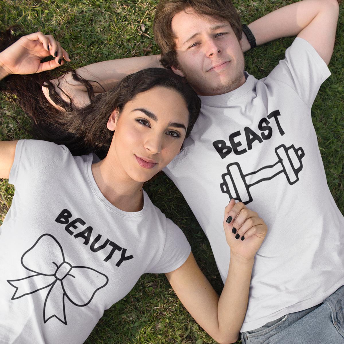 Set de două tricouri albe personalizate pentru cuplu, model 'Beauty and Beast' cu grafică fitness: tricou damă cu mesaj 'Beauty' și fundiță neagră, tricou bărbat cu mesaj 'Beast' și simbol halteră, purtate de un cuplu într-un decor natural.