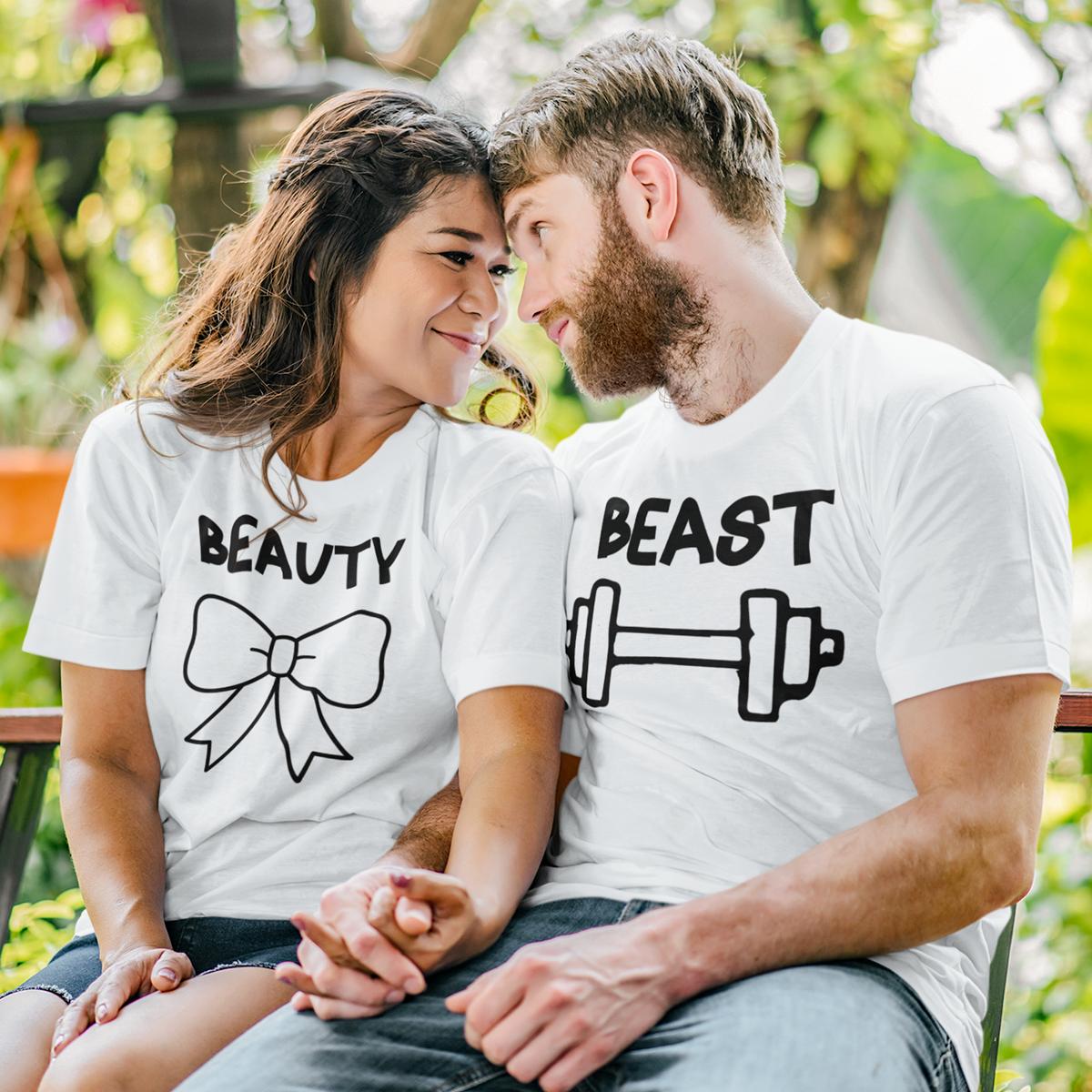 Set de două tricouri albe personalizate pentru cuplu, model 'Beauty and Beast' cu grafică fitness: tricou damă cu mesaj 'Beauty' și fundiță neagră, tricou bărbat cu mesaj 'Beast' și simbol halteră, purtate de un cuplu într-un decor natural.