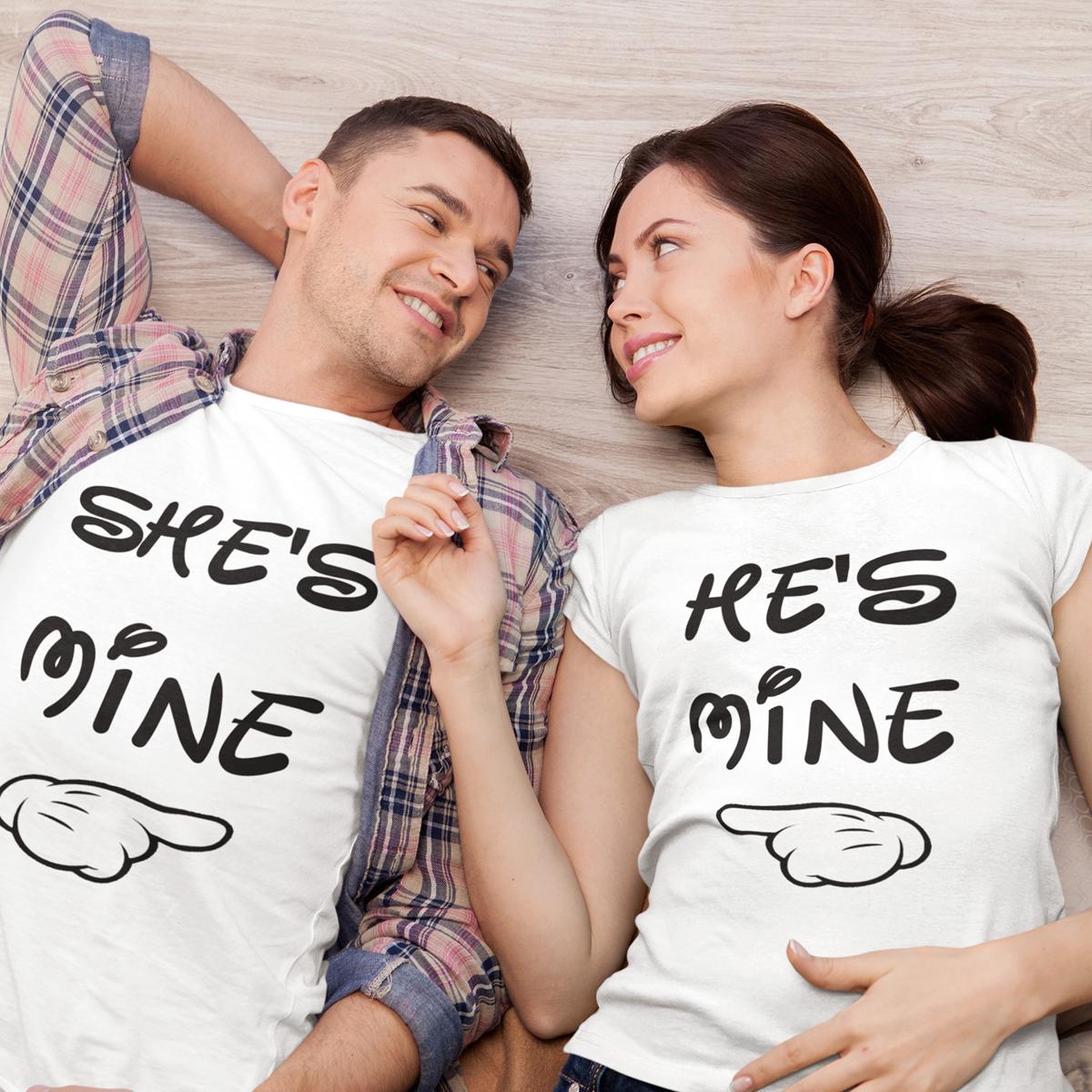 Set de două tricouri albe pentru cuplu cu mesajele albe She's Mine și He's Mine și grafică cu mână care indică partenerul, purtate de un cuplu zâmbitor