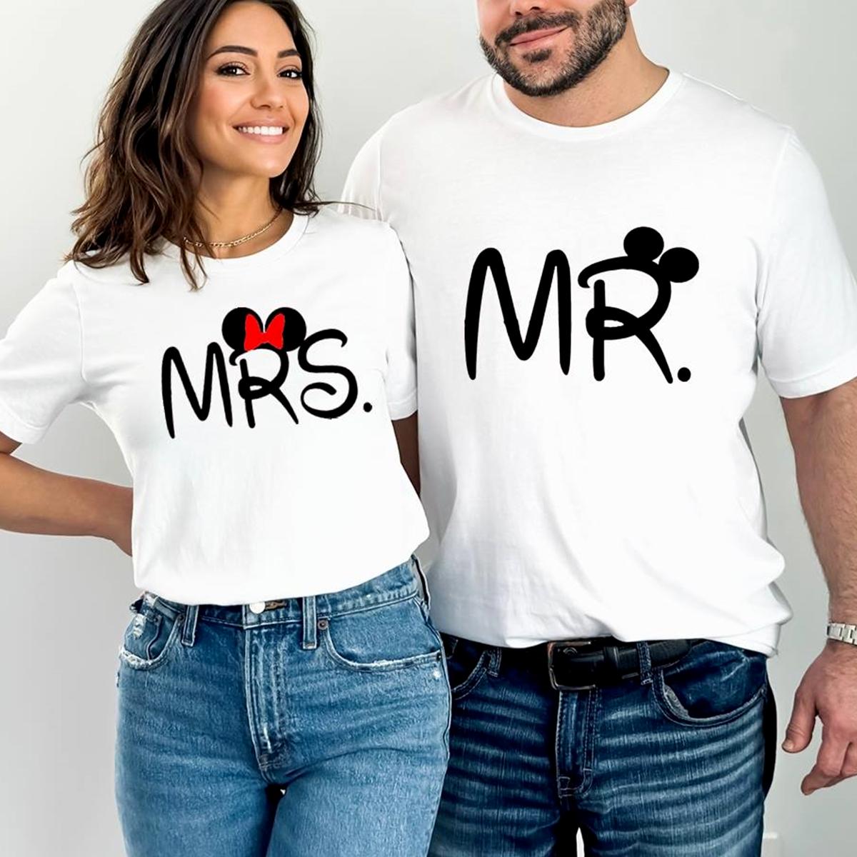 Set de două tricouri albe de bumbac pentru cuplu, imprimate cu textul „MR.” și „MRS.” într-un font negru, jucăuș. Tricoul „MRS.” este decorat cu urechi de mouse și o fundiță roșie, iar cel „MR.” include urechi de mouse integrate în litera R. Cuplul zâmbitor poartă tricourile cu blugi, oferind o sugestie de ținută casual pentru vacanțe sau ședințe foto.